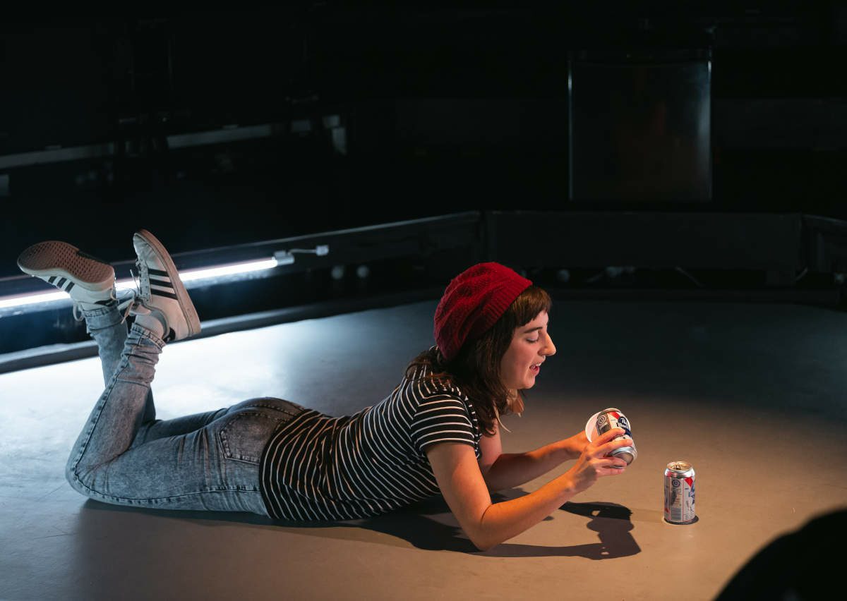Un jeune femme avec une tuque étendue par terre, les jambes croisées, en train de verser une bière dans un verre en plastique rouge