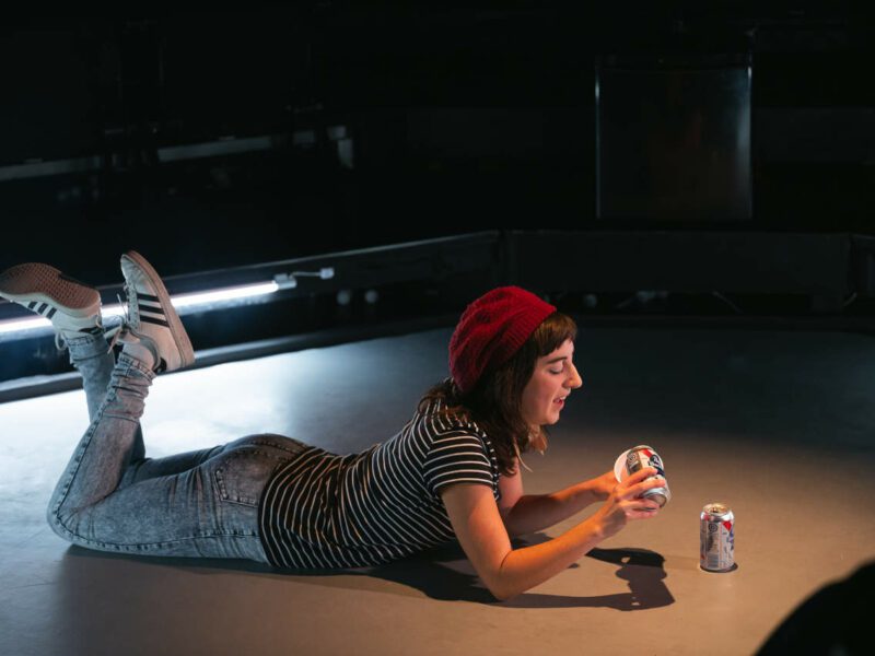Un jeune femme avec une tuque étendue par terre, les jambes croisées, en train de verser une bière dans un verre en plastique rouge