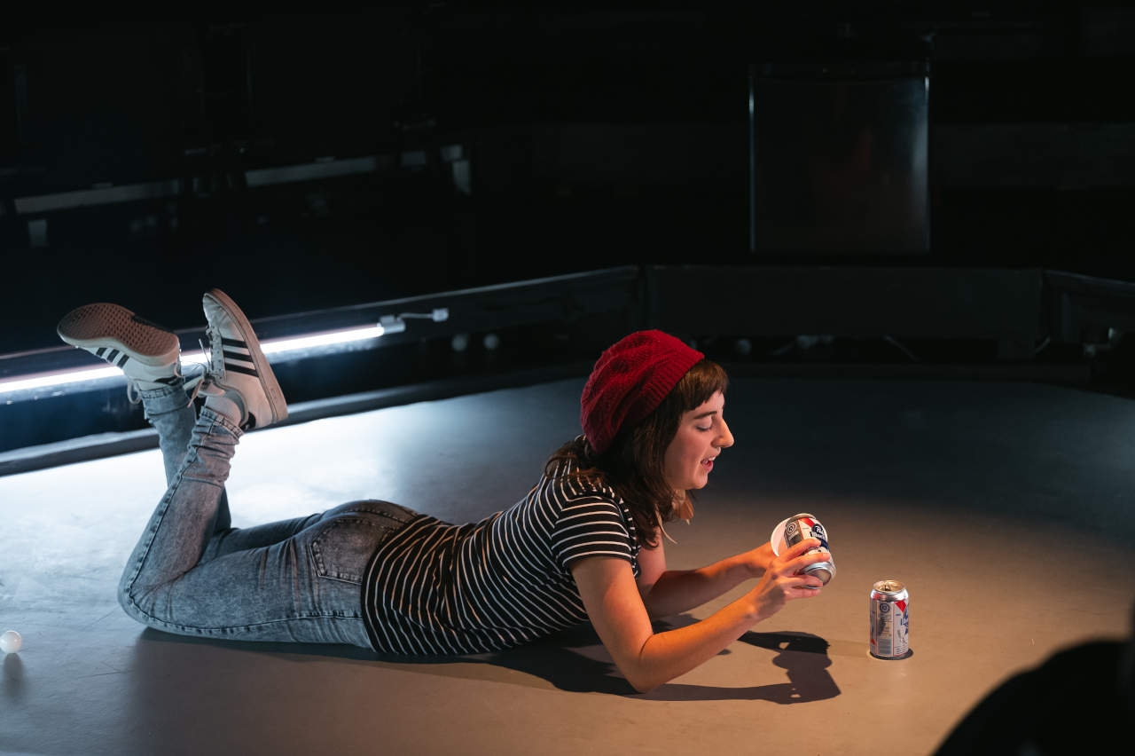 Un jeune femme avec une tuque étendue par terre, les jambes croisées, en train de verser une bière dans un verre en plastique rouge